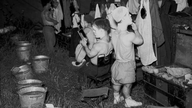 Circus performers put on makeup in a dressing area set up in a tent. The no-frills set-up includes pails of water, camp stools and trunks of clothing
